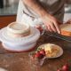 Hands preparing a layered cake with frosting, strawberries, and butter in a cozy kitchen setting.